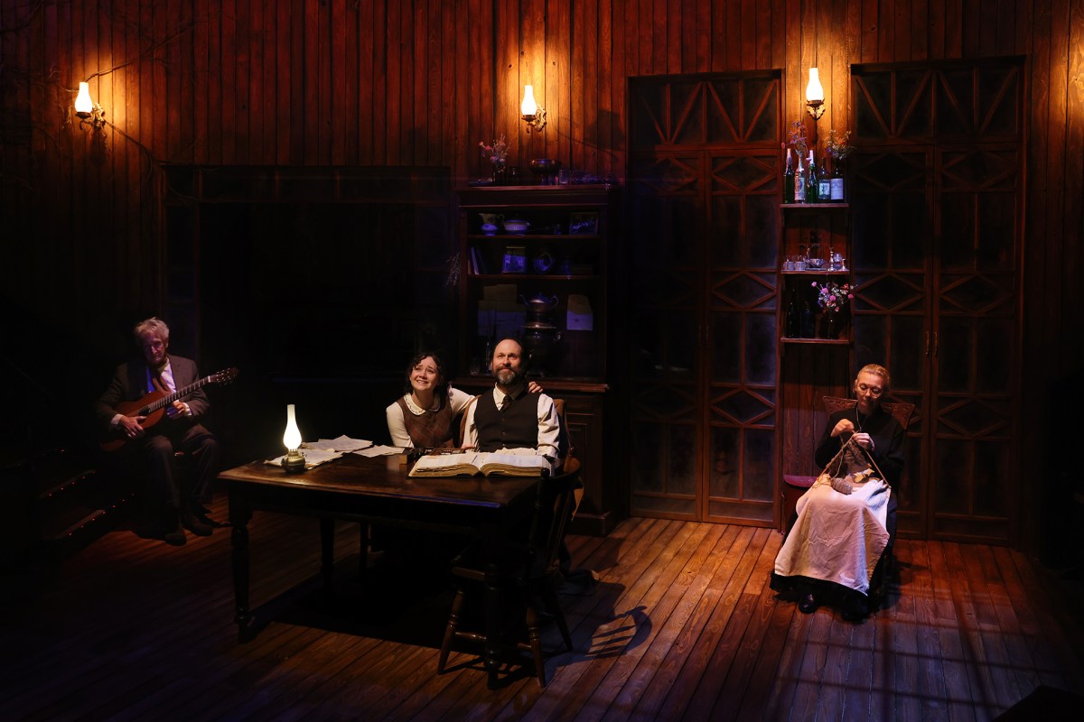 A darkened stage lit up by three small lamps. A middle-aged man and a woman are seated centrally at a table. To the left is an older man with a guitar, on the right and older woman knitting.