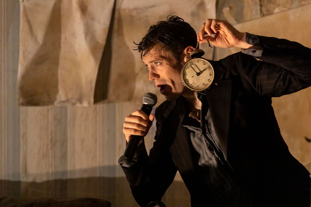 Dancer/choreographer Luke Murphy, looking sweaty and slightly dishevelled, holds an old-fashioned alarm clock up to one ear, in a scene from the dance-theatre production Volcano. He wears a dark suit jacket over a grey coat, and stands in front of a background of dingy wallpaper to which sheets of blank paper are pinned.