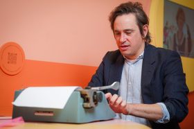 A brunette man with a light blue shirt, a dark blue jacket is typing on a blue typewriter. Trent Dalton.