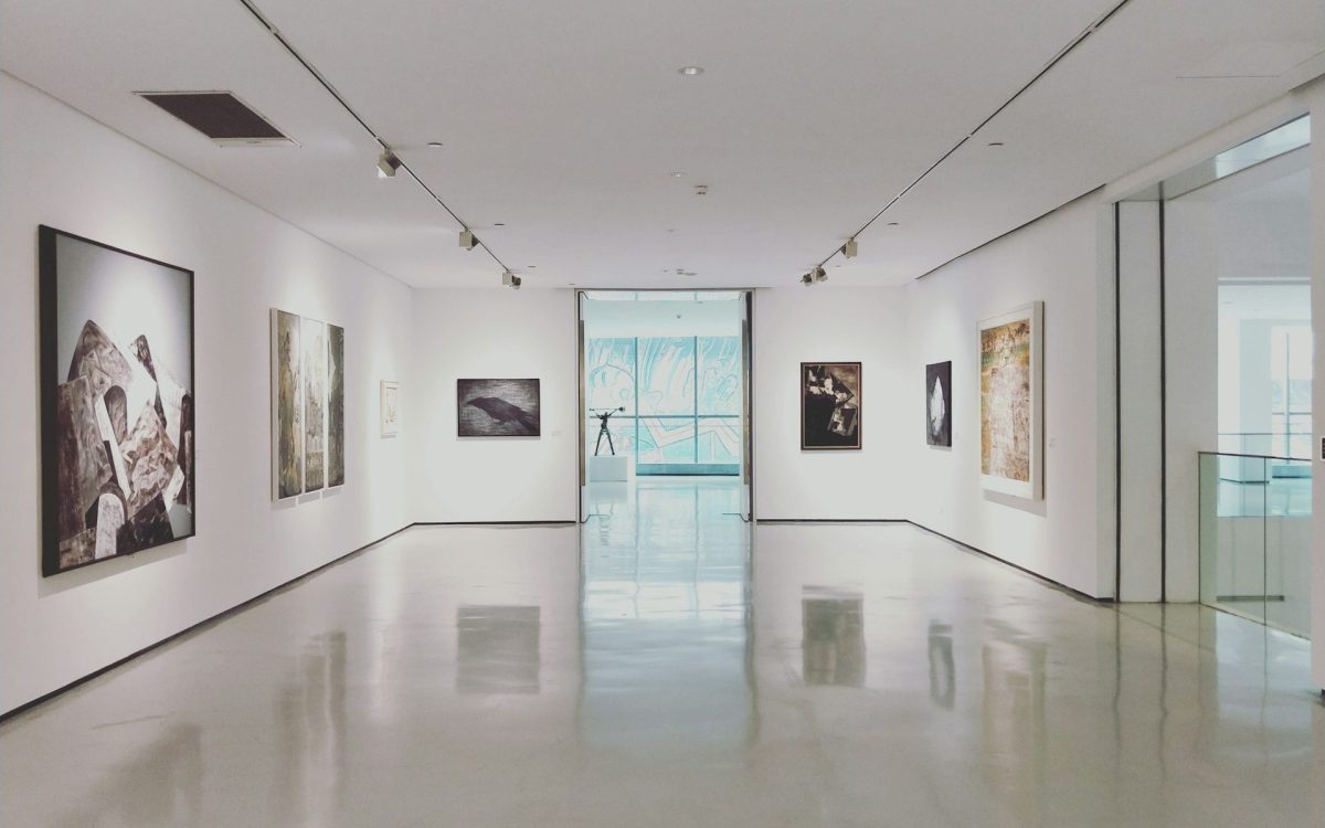 Steps to opening an art gallery as a business. Photo of a light-filled gallery space with white walls, showing a series of paintings with subdued grey and brown tones.