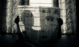 An artfully shot black and white photograph or a person reading a newspaper in front of a window. They hold thew newspaper up so that we cannot see their face, but their head is silhouetted by sunlight shining through the page.