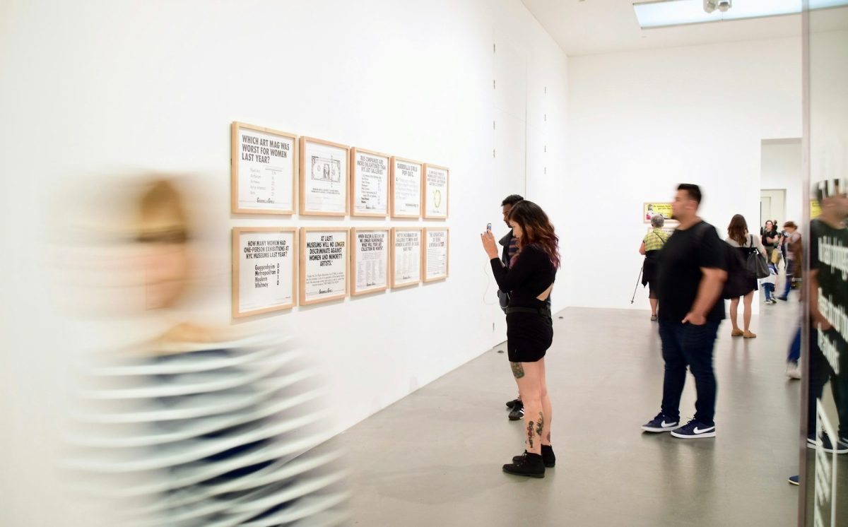 Steps to opening an art gallery as a business. Photo of a small crowd inside a contemporary gallery with white walls. A figure at the centre of the photo is standing in front of a series of 10 artworks.