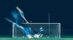 A deliberately blurred photograph of a fast-moving soccer player and goalie, the latter of whom is defending the goal from an incoming football during training.