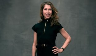 A white woman with should-length hair smiles at the camera. She wears a short-sleeved black dress and rests her left hand on her hip.