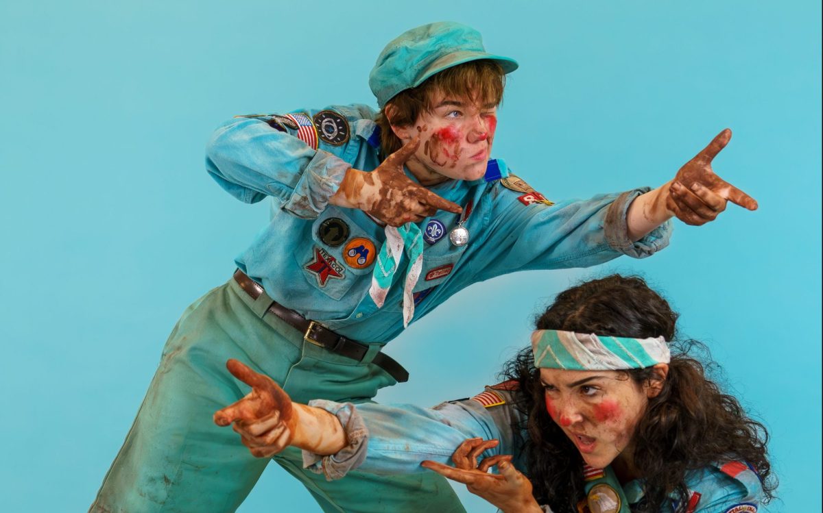 Two men with fake blood and mud on their faces and clothing and in blue scouts uniform are miming holding up guns.