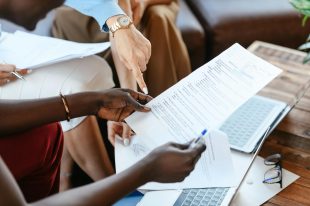 confidentiality: a photograph of hands and fingers pointing at a contract someone is holding, with papers and a laptop on a desk behind them.