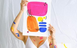 A woman holding a colourful abstract artwork that blocks out her face with colourful squiggles on her body. She is standing against a white backdrop.