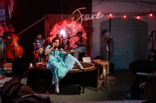 A woman in a light blue dress and silver boots is holding onto a mop. she is sitting on a table, A band is behind her.