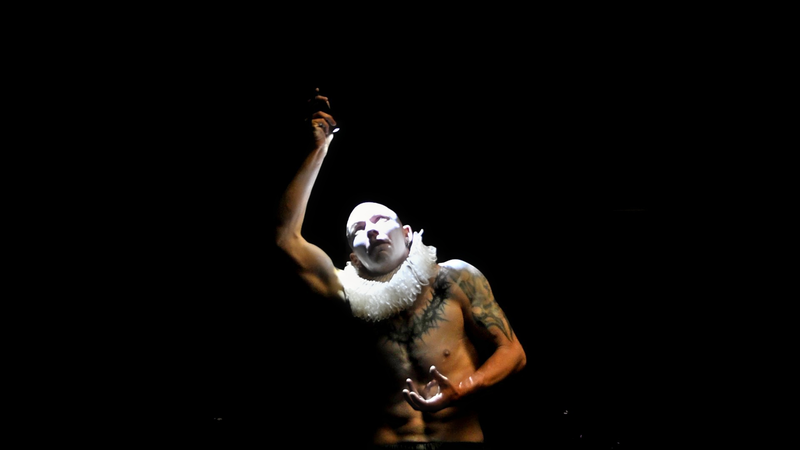 A bare-chested man is spot lit. He has tattoos on his chest and a while ruff around his neck. He has white make up on his face. His right arm is raised.