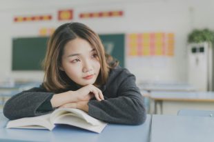 Young Asian woman is sitting at a desk with a book open in front of her but gazing off to the right as if lost in thought. Romantasy.