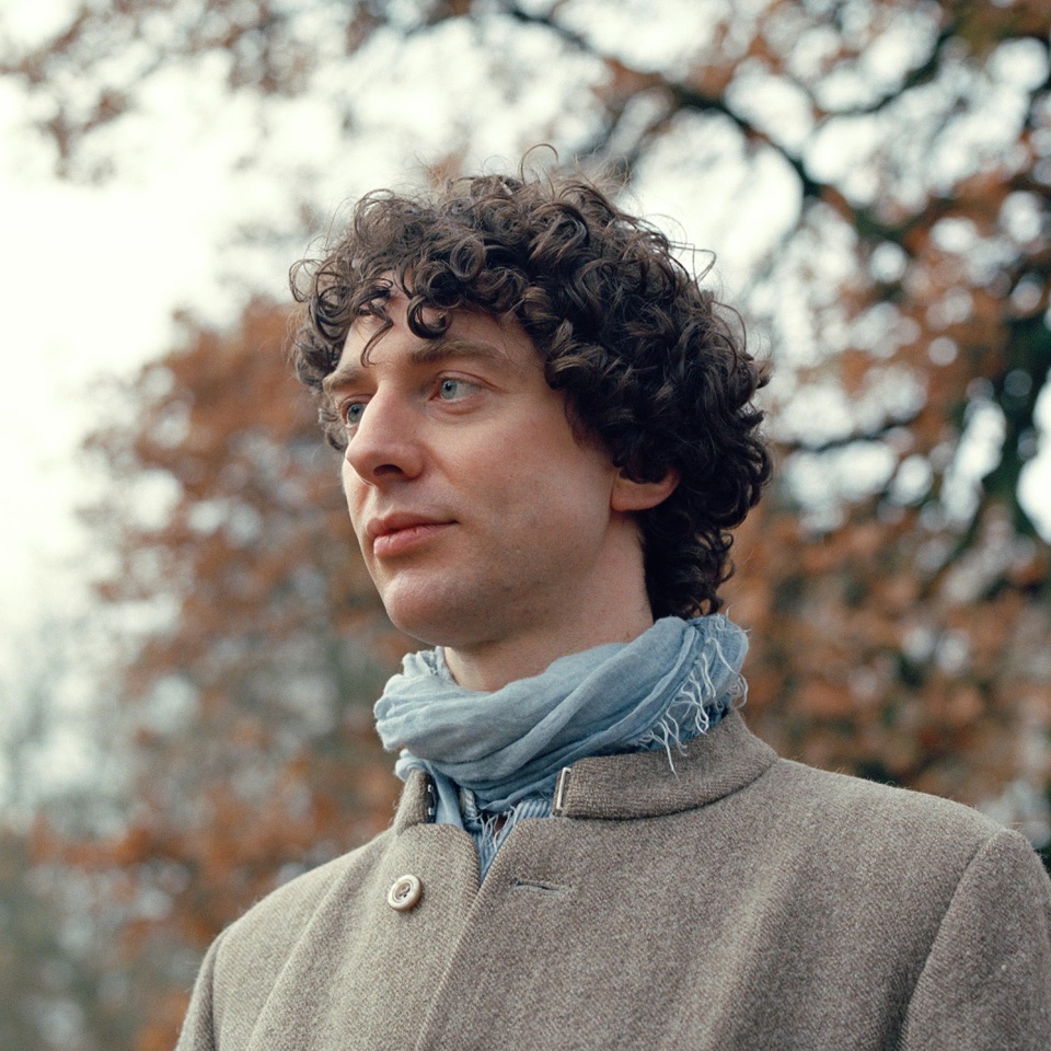 Fungi Futures. Merlin Sheldrake is a fungi expert, pictured outdoors in a light brown jacket and blue scarf. He has curly hair and is looking to the left of frame.