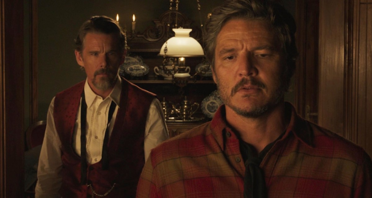 Two aging men in old-fashioned clothes stand in a dimly lit room, evocing a traditional Western movie stand-off but their expressions and positioning hint at queer undercurrents to their relationship.