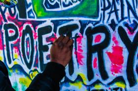 A hand is painting the word 'Poetry' in back on the wall of bright, colourful graffiti.