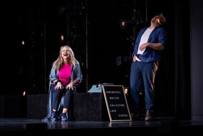 On a dark stage a blonde woman is seated and a man is standing. They are both dressed casually. There is a board between them that says 'Sentimental writer collecting love stories.'