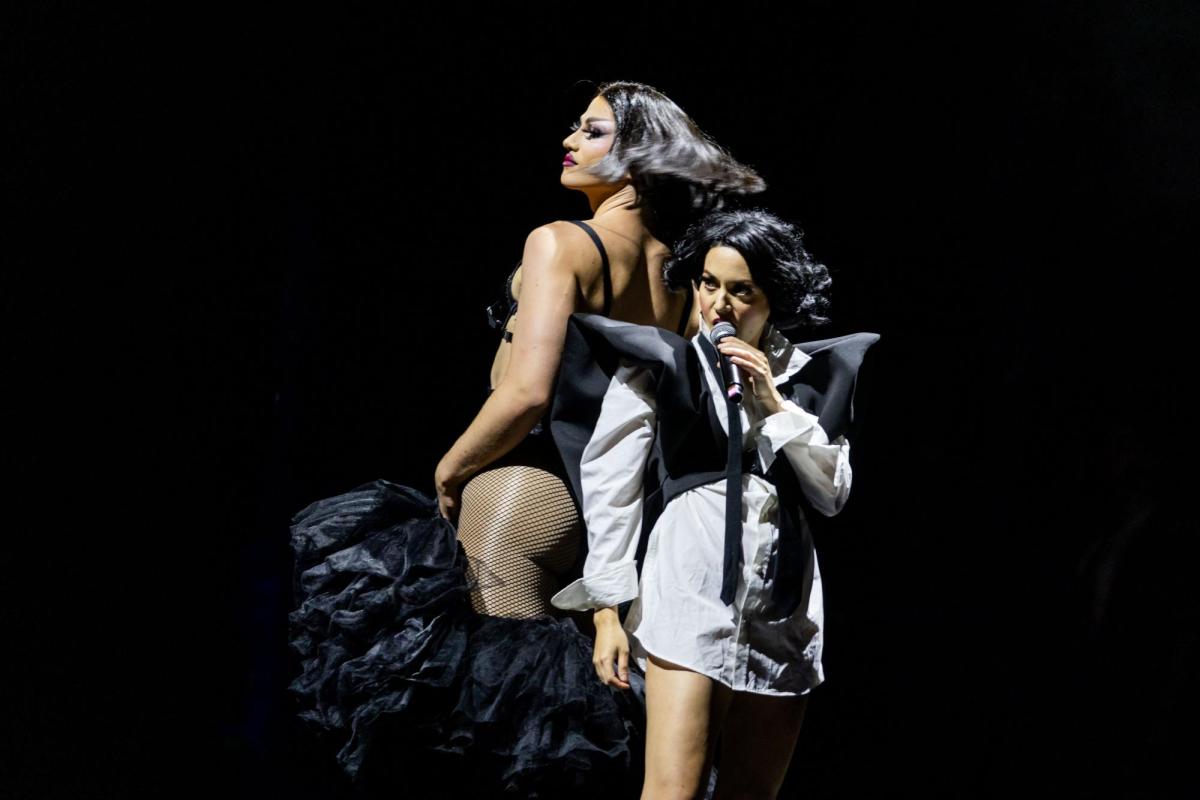 A woman, Sahara Beck, wearing a white and black outfit is standing with a microphone. A drag queen in a black leotard, is standing beside her, back to back.