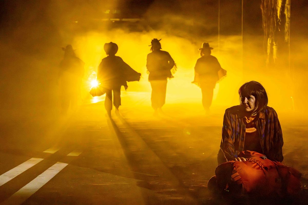 Four figures dressed like cowboys emerge from a yellow-lit haze. A woman kneels in the foreground over a backpack. This is a scene from the 2024 production of NORPA's 'Wildskin'. Photo: Kurt Petersen.