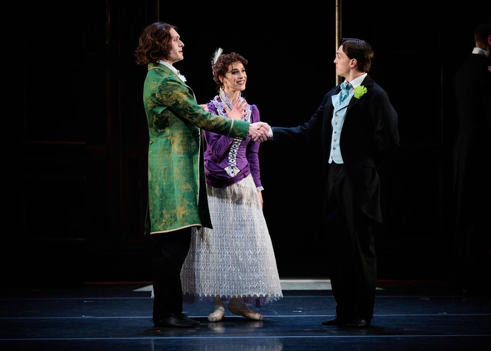 Callum Linnane as Oscar Wilde, Sharni Spencer as Constance Wilde, and Joseph Caley as Robbie Ross in The Australian Ballet's 'Oscar'. A long-haired man in a long green coat shakes the hand of a younger man who wears a green carnation in the lapel of his dark blue jacket. A smiling young woman stands between them, welcoming the younger man into their home.