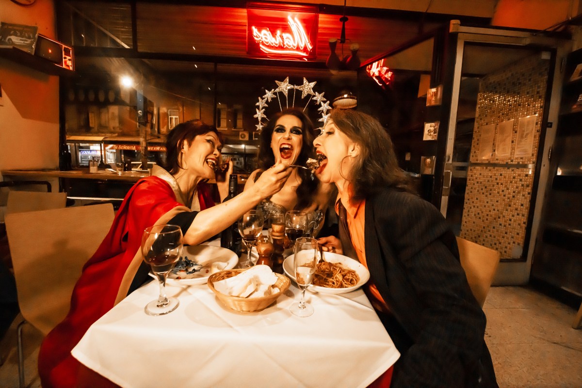 Three exotic cabaret performers feed each other extravagantly in a red hued restaurant. 