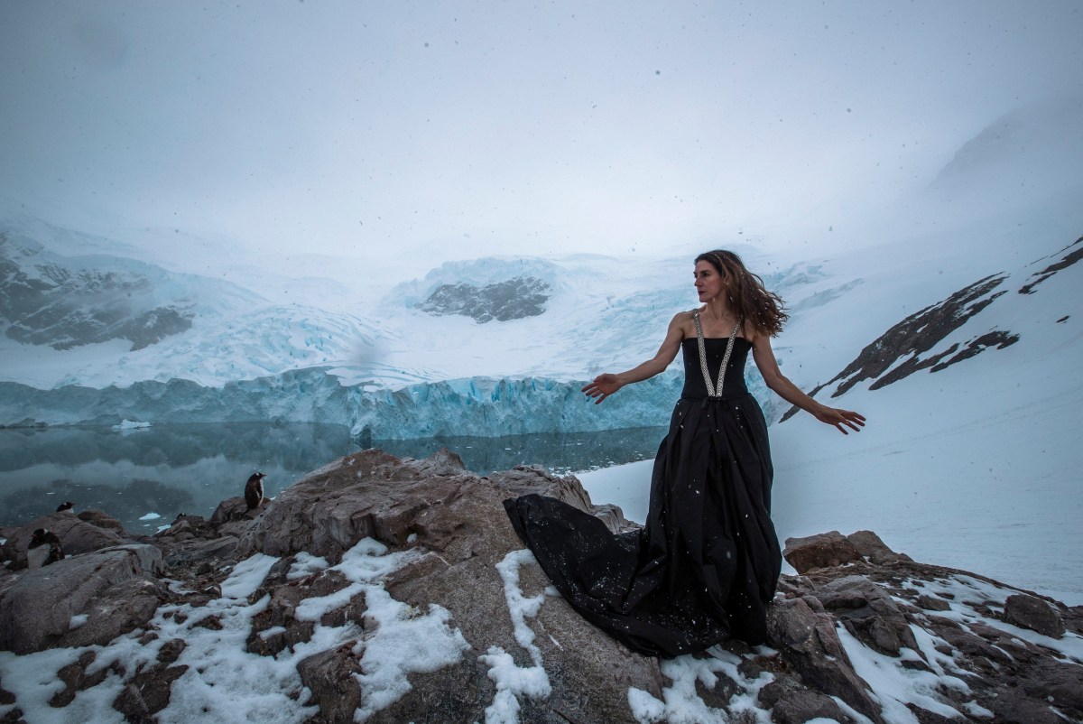 A middle aged woman in a black ball gown stands in the Antarctic wilderness with her arms stretched out to the side and looking to her right. Moira Finucane