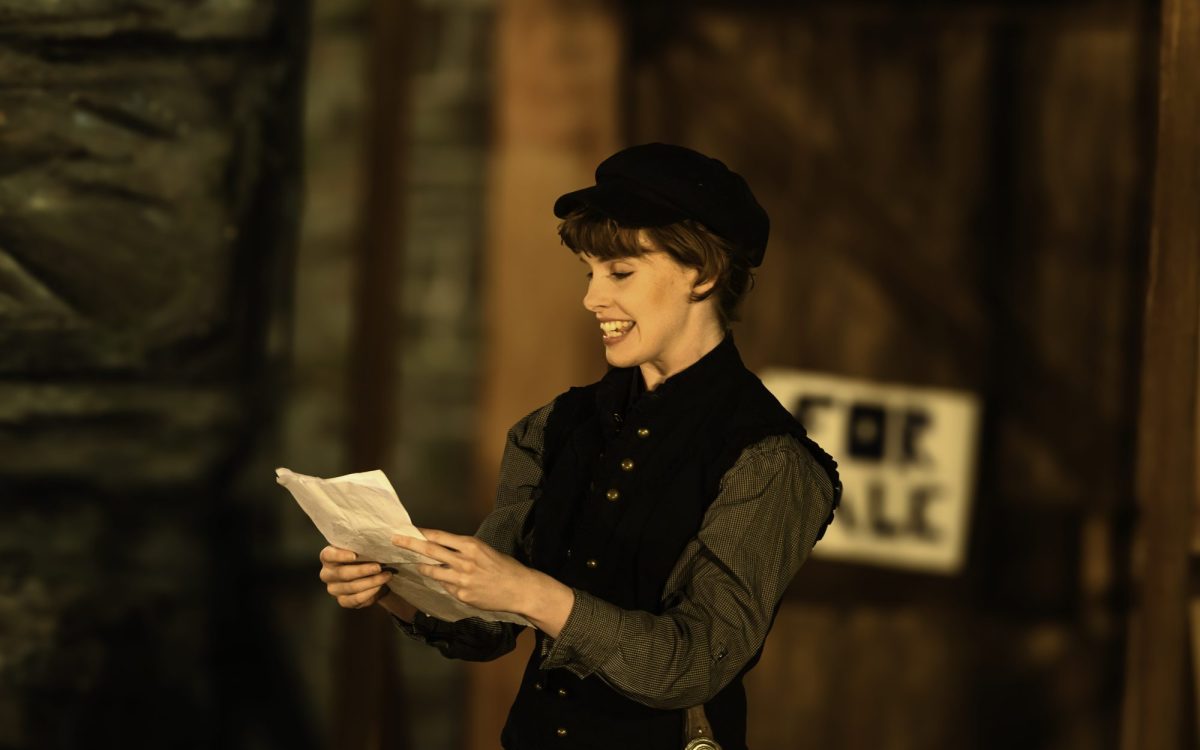 Eyse Phelan wearing a cap, vest and shirt is reading out from a piece of paper. As You Like It by TheatreiNQ