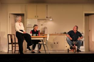 A kitchen scene. Two women are on the left at a table, one seated, another perched on the table. A man is on the right, riding a tiny child's tricycle. The Children.