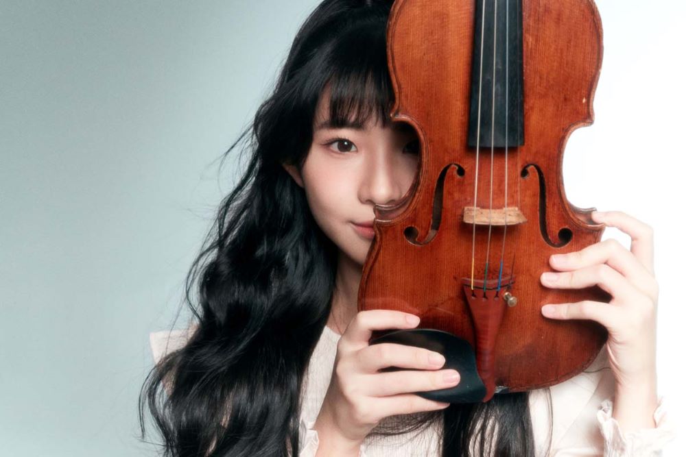 17-year-old violin sensation Chloe Chua joins the Singapore Symphony Orchestra in a concert presented by the MSO in 2025. A young Singaporean woman holds her violin close to her face and peers around it at the camera.