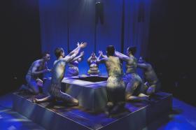 Seven dancers in 'Dredge' kneel around a water hole structure. Their hands are raised. The lighting is an eerie blue.