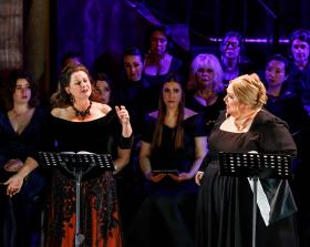 Two leading women opera singers (Deborah Humble and Helena Dix) in black with a chorus on singers in the background. Puccini.