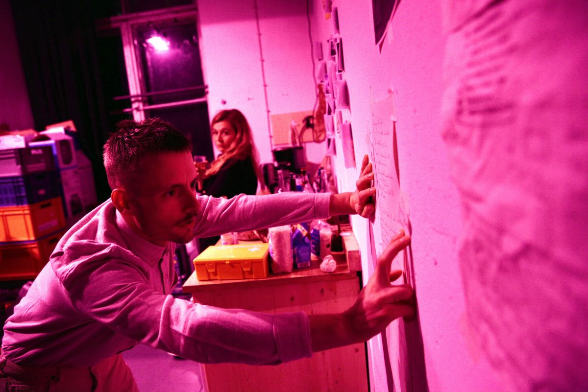 in a rose coloured scene a man is looking worried with both hands on a wall looking at plans stuck there. A woman behind seated looks on. Disaster.