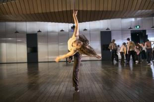 A dancer in 'The Hearth' with long brown hair is balancing on one leg in the centre of the room. Other dancers are on the right side.