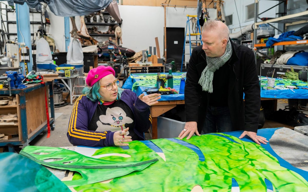 2025 Createability Internship Program for creative practitioners with disability or who are d/Deaf. Image: Past Createability participant Holly Jane Cohle and Scott Andrew. A vibrant photo of Holly and Scott in discussion in front of a green and blue art piece laid out in front of them. Holly is sitting, wearing a pink beret with blue hair, a cartoon tshirt and a stripped jacket. Scott is standing and looking at holly, wearing a grey scarf and black jacket. They appear to be inside a studio or warehouse.