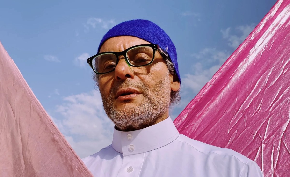 Older man with grey whiskers and glasses wearing white shirt and blue cap against blue sky and pink fabric. Designer Hassan Hajjaj