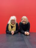 Two grey haired women sit behind a long table with their hands clasped in front of them and a red backdrop behind. On the left Suzie Dee has a messy bun, glasses and a light brown scarf. On the right Patricia Cornelius wears a black jumper.