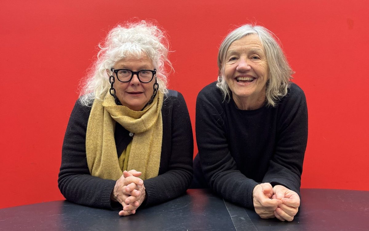 Two grey haired women sit behind a long table with their hands clasped in front of them and a red backdrop behind. On the left Susie Dee has a messy bun, glasses and a light brown scarf. On the right Patricia Cornelius wears a black jumper.