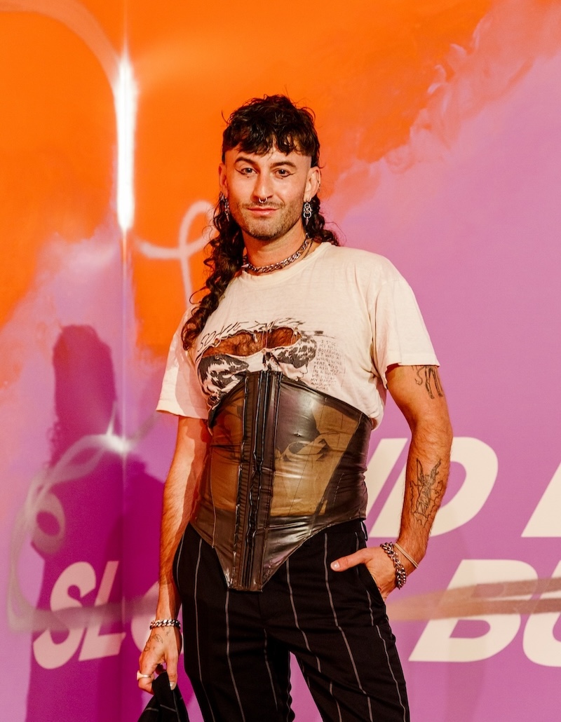 Arty man with long hair wearing a t-shirt with corsete and black pants. Curator Jake Treacy