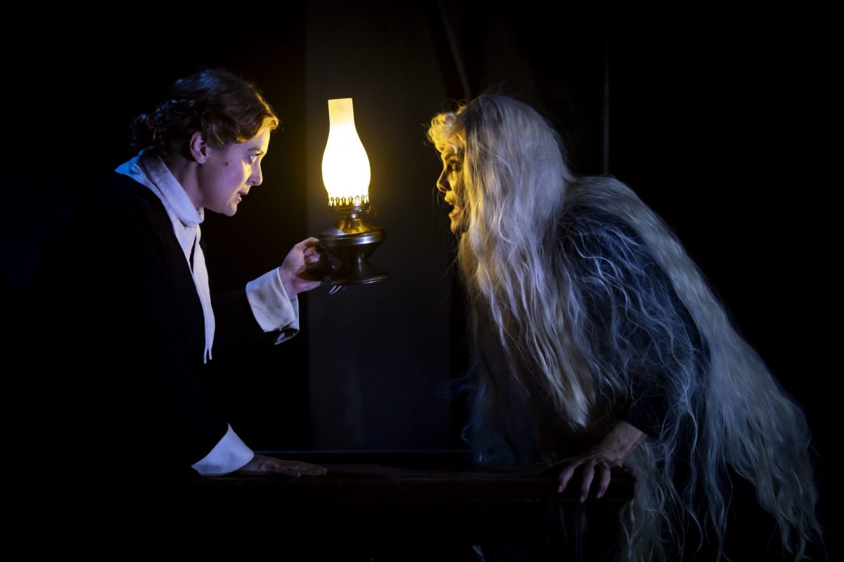 Two woman are enshrouded in darkness. The one on the left, who has short dark hair, is holding a lamp. The other one has long white hair. The Turn of the Screw by Hayes Theatre.