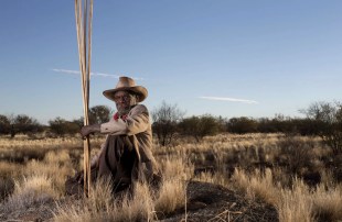 Aboriginal man wearing hat and holding spears in country landscape.