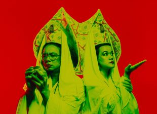 Dressed in yellow and green robes with ornate headwear, two Asian-Australian artists, Joe Paradise Lui (left) and Merlynn Tong (right), pose against a red background. Promotional image for the 2025 MTC production 'Legends (of the Golden Arches)'. MTC 2025 season.