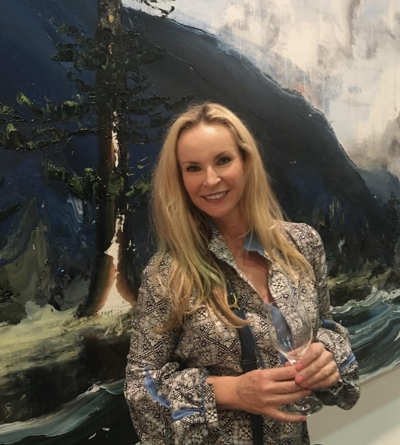 Woman with long blonde hair wearing patterned shirt standing in front of a painting. Curator Lizzy Galloway.