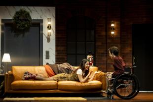 A young woman, Mabel Li, is lying on her belly on a couch. She is talking to a young man, Oli Pizzey Stratford, who is in a wheelchair, in a scene from MTC's 'Cost of Living'.