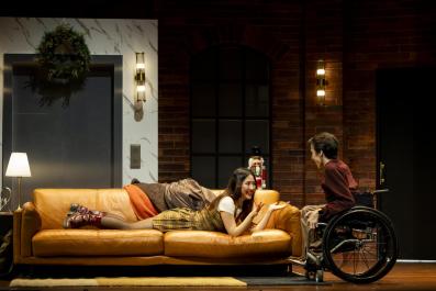 A young woman, Mabel Li, is lying on her belly on a couch. She is talking to a young man, Oli Pizzey Stratford, who is in a wheelchair, in a scene from MTC's 'Cost of Living'.