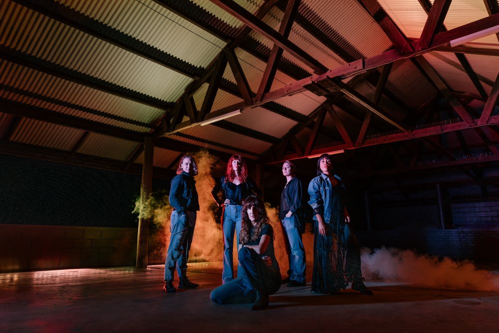 The 2024 cast of NORPA's production Wildskin stand beneath the wooden beams and iron roof of the Lismore Showground pavilion. Four of the women are standing, one is half-crouched on the ground before them. All five women wear jeans and denim jackets or jeans and black shirts. Smoke billows dramatically behind them.