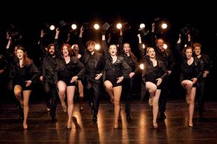 A group of dancers, all in black, the women in stockings and the men in trousers, all holding top hats in the air, in the production of 'A Chorus Line.'