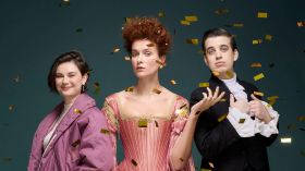 A publicity image for Belvoir's 2025 adaptation of Virginia Woolf's Orlando. A dark-haired woman in a purple jacket, a red-haired woman in a period dress, and a dark-haired young man in a ruffled shirt and frock coat look towards the camera. Golden glitter showers down around them.