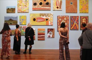 A picture of around 10 different Aboriginal art paintings hanging on a gallery wall showing four gallery attendees of different ages and genders looking at them.