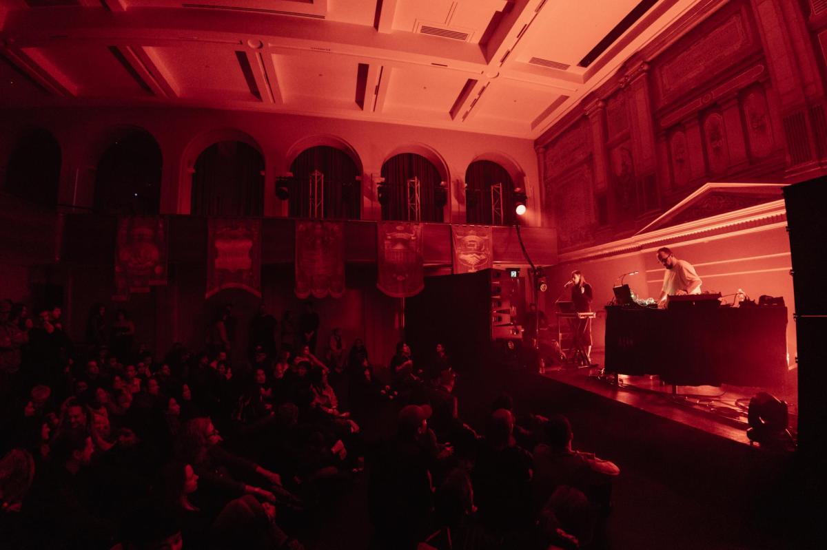 A concert scene. Audience on the left, stage on the right. The lighting is red.