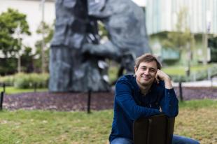 Trent Dalton, a smiling man with brown hair ins sitting in a garden in front of a grey sculpture.