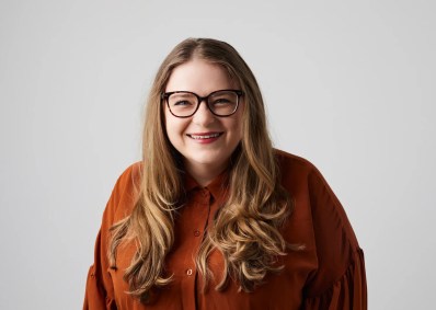 A long haired young woman in glasses wearing a brown shirt and smiling at the camera.