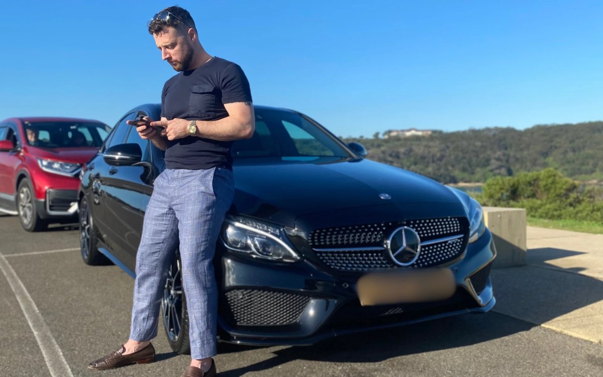 A man in light trousers and a grey T shirt leans against the bonnet of a car looking at a phone