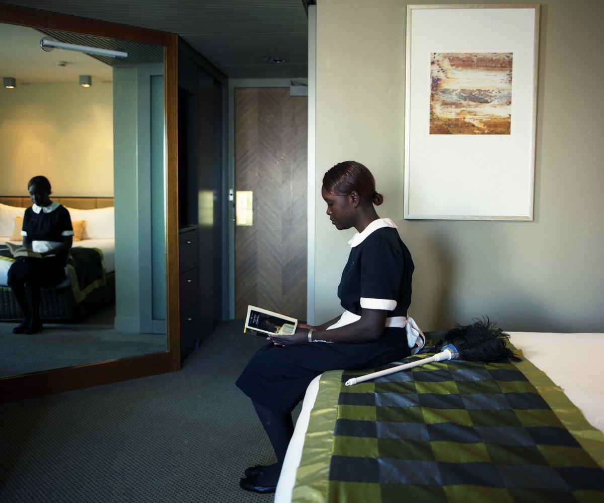 Anne Zahalka. Hotel Suite series. A black maid sits in a hotel room on the made up bed, reading a Penguin classic novel. She is reflected in the mirror and has a feather duster next to her on the bed.